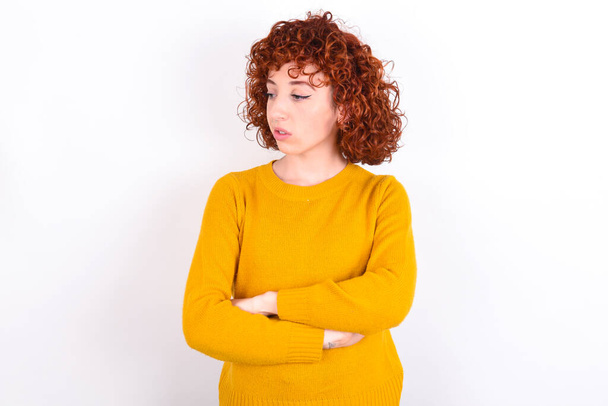 Недовольная рыжая девушка в жёлтом свитере на белом фоне с плохим настроем, руки скрещены в сторону. Отрицательные человеческие эмоции. - Фото, изображение