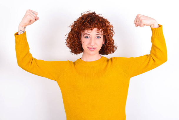 Waist up shot of young redhead girl wearing yellow sweater over white background   raises arms to show muscles feels confident in victory, looks strong and independent, smiles positively at camera. Sport concept. - Photo, Image