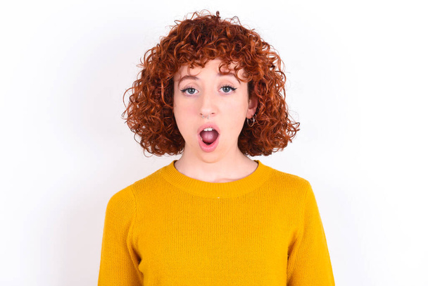 Emotional attractive young redhead girl wearing yellow sweater over white background   with opened mouth expresses great surprise and fright, stares at camera. Unexpected shocking news and human reaction. - Photo, Image