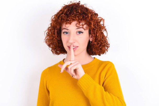 Sorridente giovane rossa ragazza che indossa maglione giallo su sfondo bianco fa un gesto di zittire, tiene il dito indice sulle labbra nasconde segreto. Sii muto, per favore.. - Foto, immagini