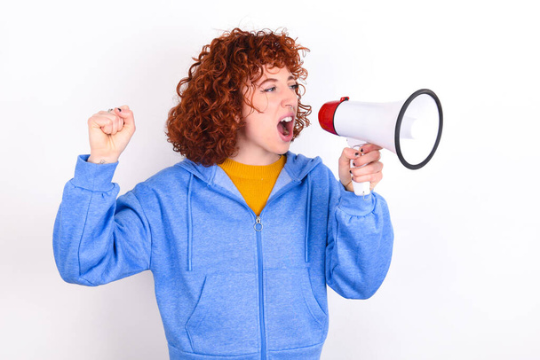 fiatal vörös hajú lány visel kék kabát fehér háttér kommunikál kiabál hangosan kezében egy megafon, kifejezve a siker és a pozitív koncepció, ötlet marketing vagy értékesítés. - Fotó, kép
