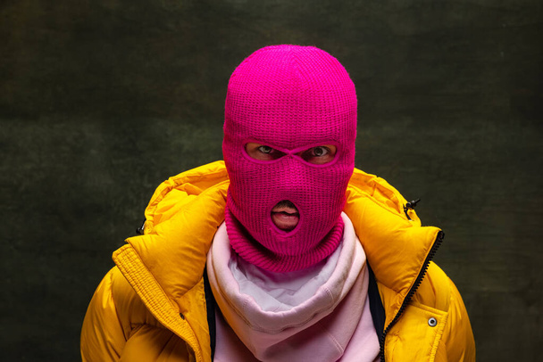 Studio shot of young anonymous man wearing pink balaclava and yellow down jacket, coat isolated on dark vintage background. Concept of safety, art, fashion - Photo, image