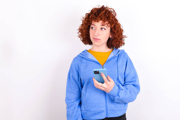 молода руда дівчина в синій куртці над білим тлом тримає мобільний телефон використовує високошвидкісний Інтернет і соціальні мережі мають онлайн-спілкування. Концепція сучасних технологій
 - Фото, зображення
