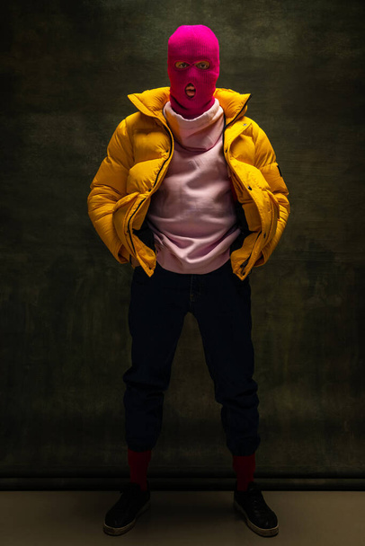 Studio shot of young anonymous man wearing pink balaclava and yellow down jacket, coat isolated on dark vintage background. Concept of safety, art, fashion - 写真・画像