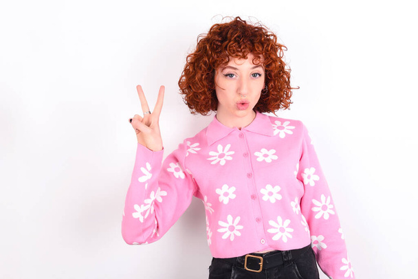 young redhead girl wearing pink floral t-shirt over white background makes peace gesture keeps lips folded shows v sign. Body language concept - Photo, Image