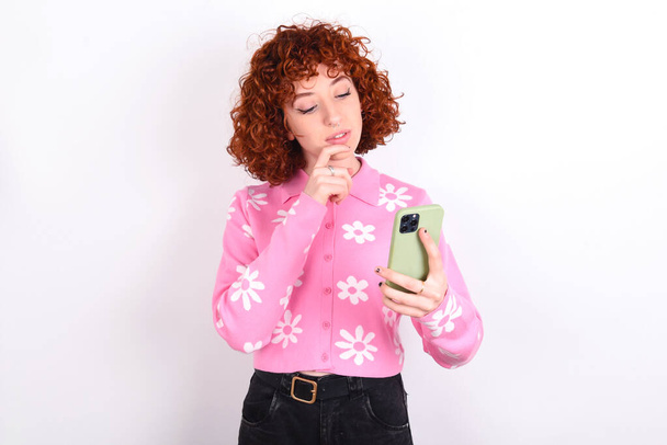 Gondolkodó boldog fiatal vörös hajú lány visel rózsaszín virágos póló felett fehér háttér tartsa meg telefonon, másolás - Fotó, kép
