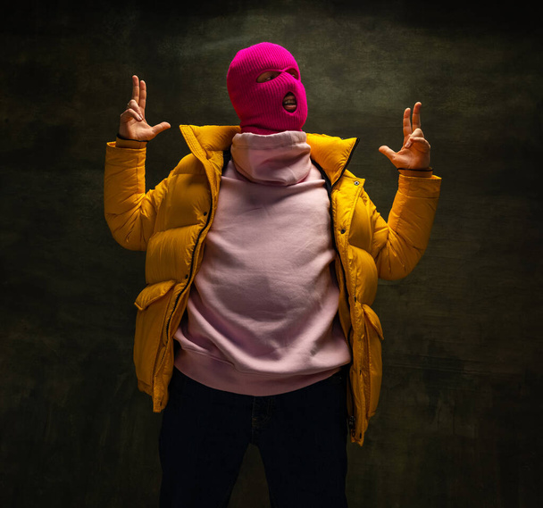 Studio shot of young anonymous man wearing pink balaclava and yellow down jacket, coat isolated on dark vintage background. Concept of safety, art, fashion - Zdjęcie, obraz