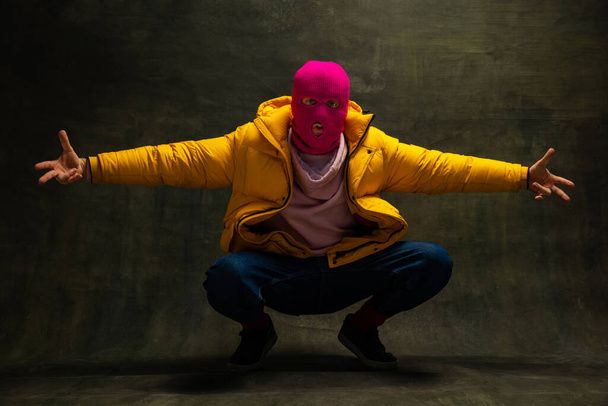 Studio shot of young anonymous man wearing pink balaclava and yellow down jacket, coat isolated on dark vintage background. Concept of safety, art, fashion - Photo, Image