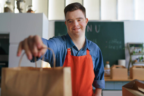Cheerful young Down Syndrome waiter holding take away order in restaurant, social inclusion concept. - Fotografie, Obrázek