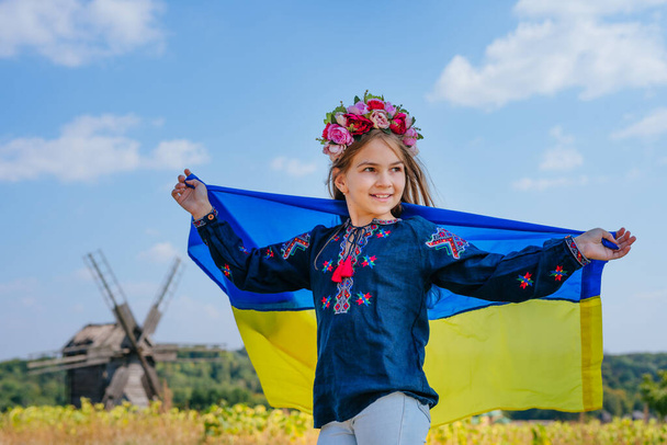 Girl wearing national clothes with embroidery and wreath holding flag of Ukraine - Photo, Image