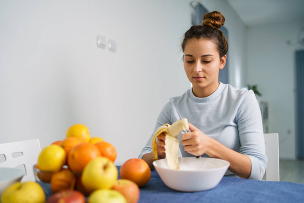 Biała młoda kobieta obierająca owoce siedząc przy stole w domu - widok z przodu na ładną dziewczynę trzymającą nóż i przygotowującą posiłek bananowy - zdrowe jedzenie przestrzeń kopiująca - Zdjęcie, obraz
