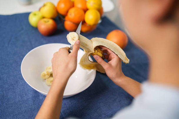 Close up on hands of unknown caucasian woman cutting banana by the table at home - back view top angle copy space - healthy eating concept - Zdjęcie, obraz