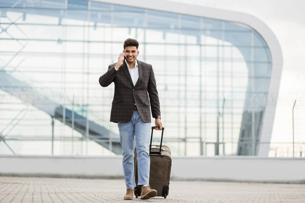 Voyageur homme d'affaires arabe parlant au téléphone, tout en marchant à l'extérieur de l'aéroport moderne, portant une valise - Photo, image