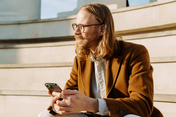 Ginger man smiling and using mobile phone while sitting on stairs outdoors - Photo, Image
