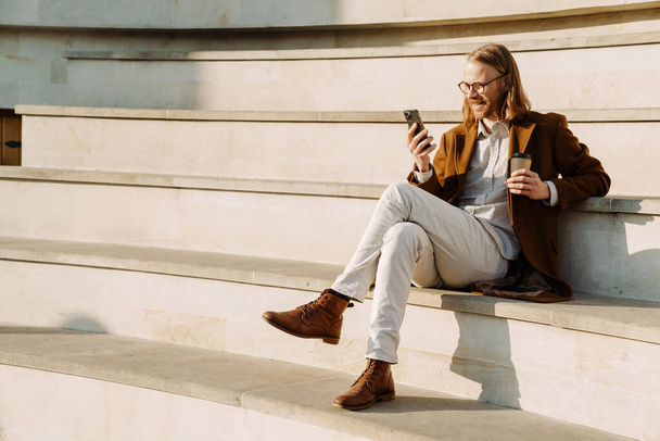 Ginger man using mobile phone and drinking coffee while sitting on stairs outdoors - Foto, afbeelding
