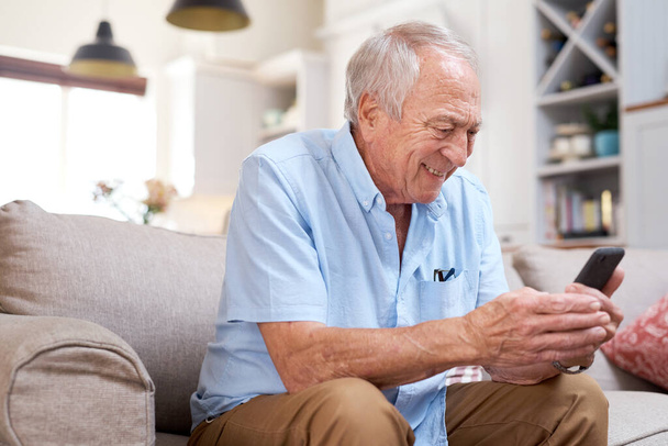 Nézzenek oda... Egy idős férfiról, aki boldognak tűnik, miközben otthon használja az okostelefonját a kanapén.. - Fotó, kép