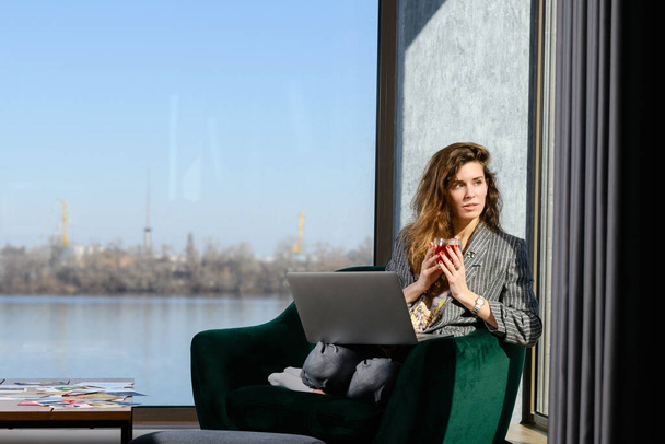 Mujer joven sentarse con el ordenador portátil y la taza de té en el acogedor interior de la casa, apartamento moderno con ventana panorámica y vista al río. - Foto, imagen