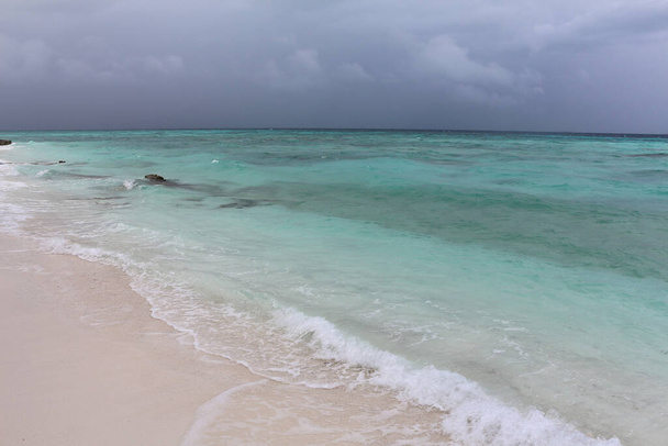 Malediwy, plaża z białym piaskiem w pobliżu turkusowego oceanu. Niebo z burzowymi chmurami nad spokojnym morzem. - Zdjęcie, obraz