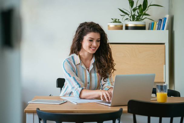 Uśmiechnięta kobieta wpisująca raport biznesowy na klawiaturze laptopa i wykonująca papierkową robotę siedząc przy biurku ze szklanką soku i tabletem cyfrowym w biurze - Zdjęcie, obraz