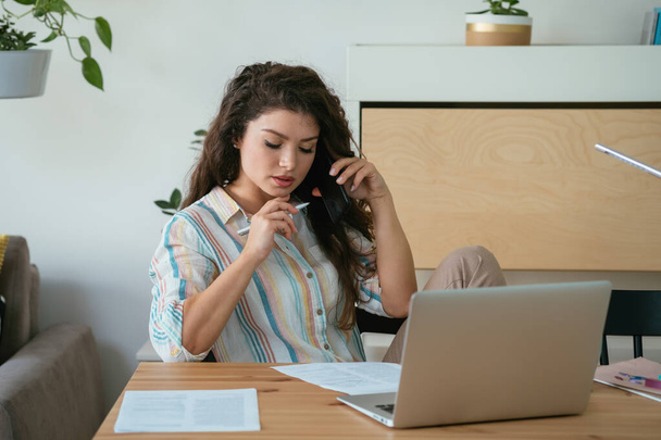 Зайнята жінка розмовляє по мобільному телефону в домашньому офісі. Серйозна бізнес-жінка розмовляє по телефону з клієнтом, сидячи за столом і працюючи над своїм ноутбуком.  - Фото, зображення