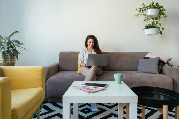 Mujer de negocios feliz usando una computadora portátil en casa. Trabajar en casa: Mujer sonriente escribiendo informe de negocios en un teclado portátil mientras está sentado en un sofá en una sala de estar. - Foto, Imagen