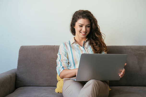 自宅でソファに座っている間、ラップトップコンピュータを使用して幸せなビジネス女性。自宅で働く:リビングルームのソファに座っている間、ノートパソコンのキーボードにビジネスレポートを入力する笑顔の女性. - 写真・画像