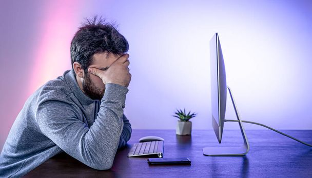 Väsynyt mies istuu tietokoneen edessä, peittää kasvonsa käsillään. - Valokuva, kuva