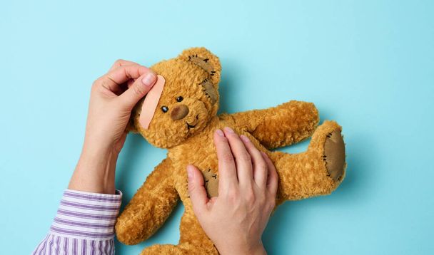 vrouwelijke hand houdt een bruine teddybeer vast en lijmt een medisch lijmpleister op een blauwe achtergrond, trambehandeling - Foto, afbeelding
