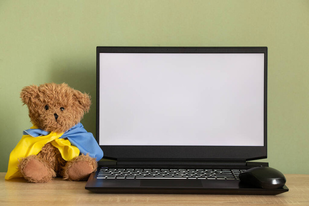 Teddy bear with flag of Ukraine near laptop with white screen on table at home, war in Ukraine, patriot, template, peace in Ukraine, laptop with white screen - Фото, зображення