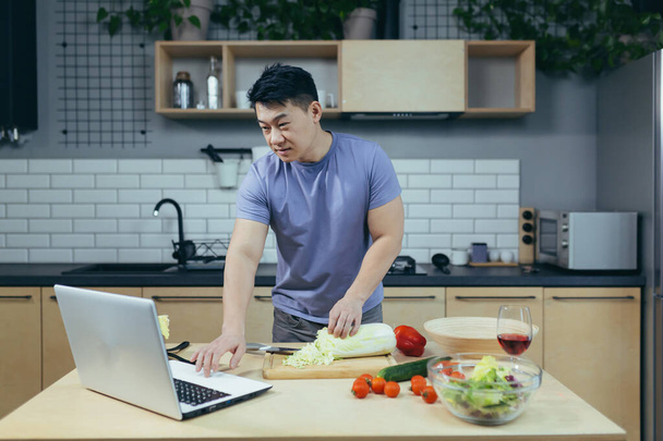 cocineros masculinos en casa y enseña en línea, cortes asiáticos verduras en la cocina, utiliza ordenador portátil para las clases de cocina en línea - Foto, Imagen