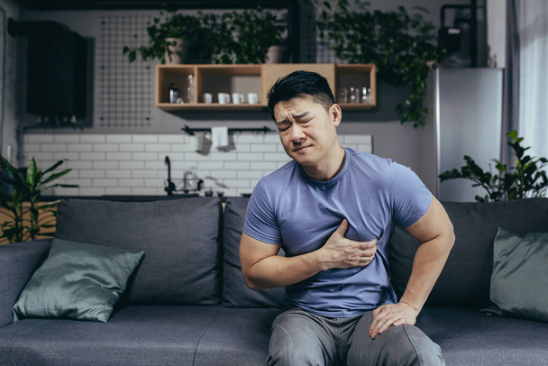 A man at home sitting on a sofa in the living room, an Asian patient has severe chest pain, heart disease - Valokuva, kuva