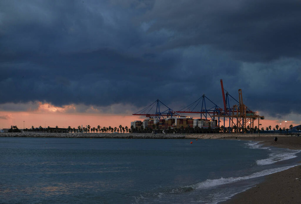 Πολύχρωμο ηλιοβασίλεμα πάνω από το λιμάνι και βιομηχανικούς γερανούς. Ακτογραμμή της παραλίας με θέα το λιμάνι της Ισπανίας. Μεταφορά και υλικοτεχνική υποδομή - Τα εμπορευματοκιβώτια της θάλασσας εκφορτώνονται από πλοία με τεράστιους γερανούς - Φωτογραφία, εικόνα