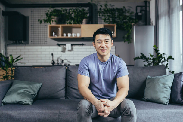 Portrait of a man at home, Asian himself smiling and looking at the camera sitting on the couch - Photo, image