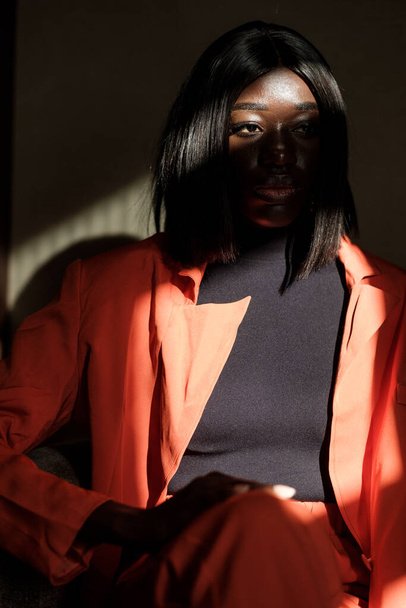 Fashion portrait of black model in shadows. She is wearing an orange jacket. - Фото, зображення