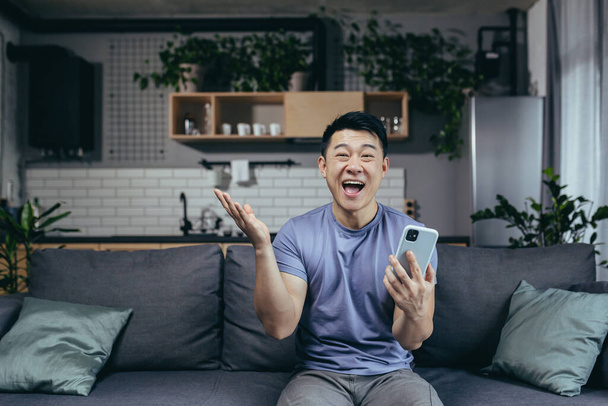 Happy man at home, successful win, won online sitting on the couch, Asian screams with joy and smiles looks at the camera, holds the phone - Photo, image