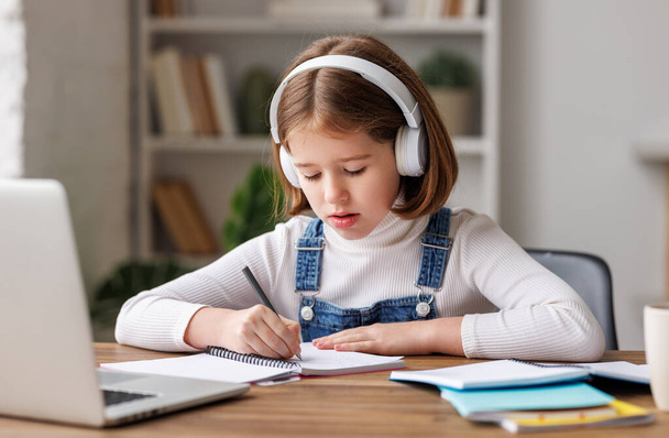 Enfant concentré en vêtements décontractés avec écouteurs sans fil assis au bureau et écrivant dans un planificateur près d'un ordinateur portable pendant la leçon en ligne le jour à la maison - Photo, image
