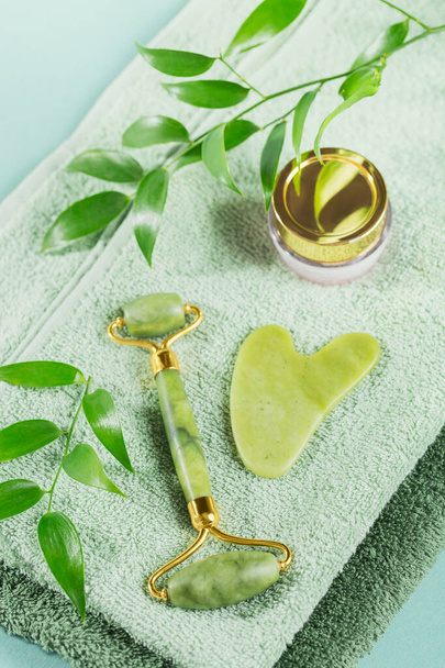 Green jade face roller and gua sha scraper with leaves on light blue background - Valokuva, kuva