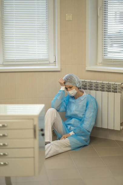 Una doctora agotada se sienta cansada en el suelo del hospital con una máscara facial y uniforme azul. - Foto, Imagen