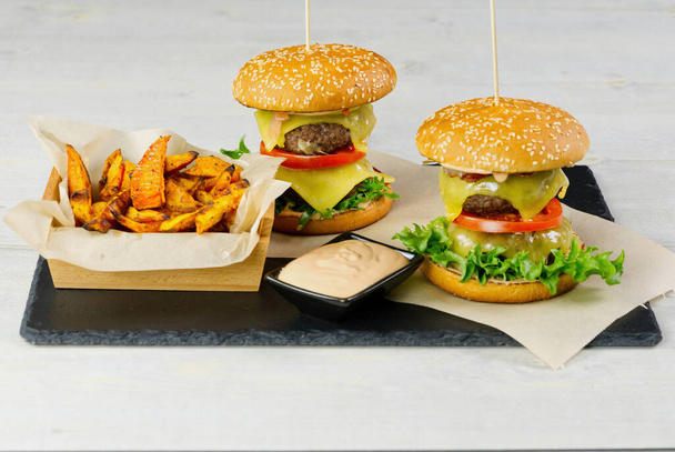 two Big burgers and sweet potato isolated on black stone plate. light wooden background - Foto, imagen