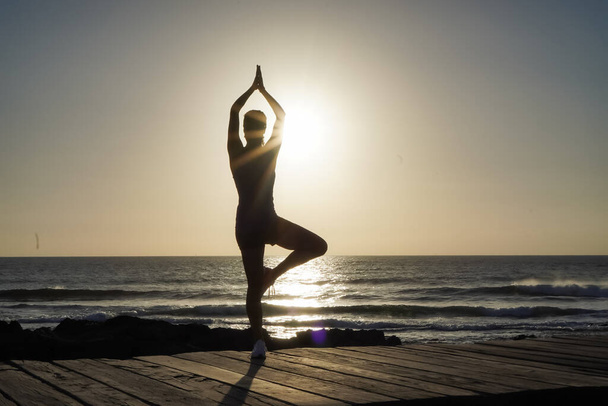 silhouette of a sportive female in namaste pose practicing yoga & meditation on beach boardwalk during sunset or sunrise  - Fotografie, Obrázek