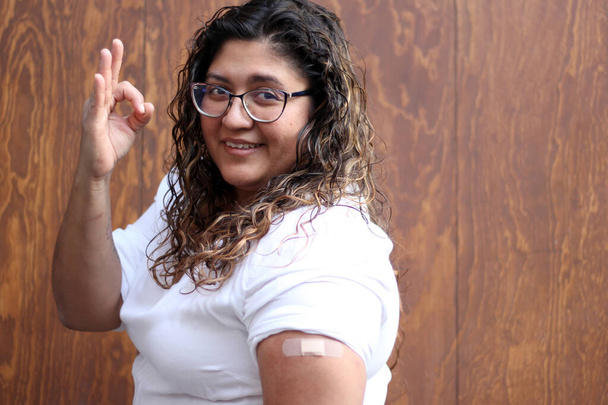 Mulher adulta latina com blusa branca cabelo ondulado e óculos mostra seu braço com uma bandagem porque ela acaba de ser vacinada contra Covid-19 na nova normalidade devido à pandemia de Coronavirus - Foto, Imagem