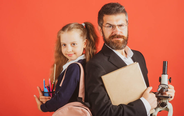 Elementary pupil with serious teacher in red studio, isolated. Portrait of funny school girl and tutor with school supplies. Tutor and student girl on red. - 写真・画像