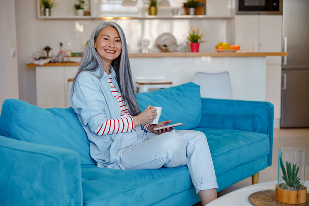 Mujer sonriente con teléfono inteligente y café sentado en el sofá en casa - Foto, Imagen