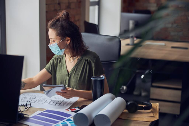 Γυναίκα διακοσμήτρια φορώντας προστατευτική μάσκα κρατώντας σημειώσεις στο σημειωματάριό της, ενώ εργάζεται σε νέο έργο, κάθεται στο τραπέζι στο γραφείο της - Φωτογραφία, εικόνα