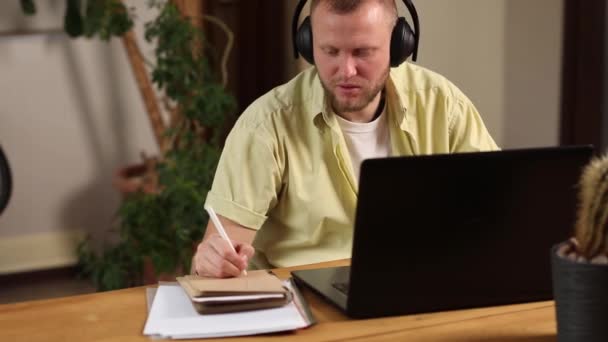 Man wearing wireless headphones watch webinar, trainning, course, lessons on laptop take notes to paper notebook at home. - Footage, Video