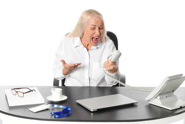 Angry mature secretary talking by phone at table on white background - Φωτογραφία, εικόνα