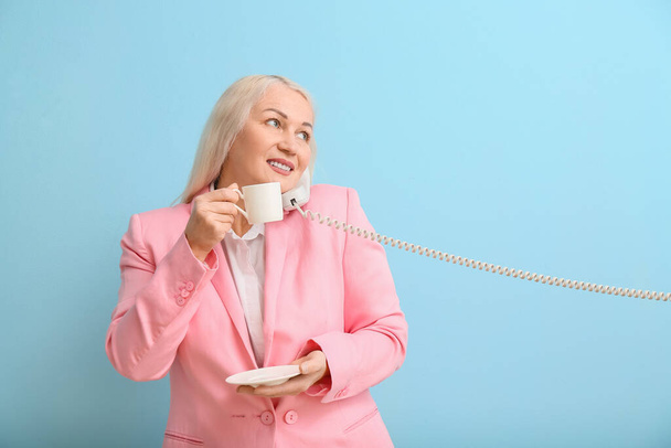 Зрелая секретарша с чашкой кофе разговаривает по телефону на синем фоне - Фото, изображение