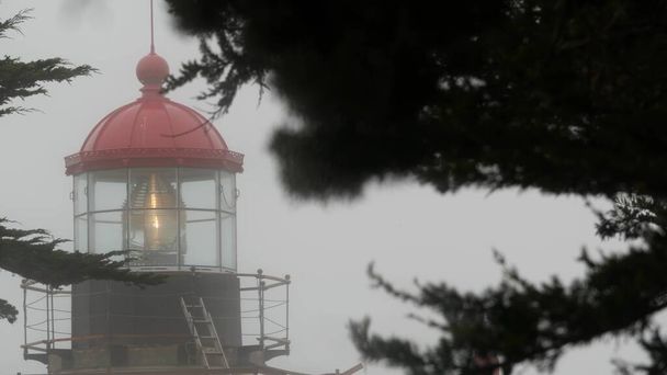Старая фрезовая линза маяка светится, туманная дождливая погода. Освещённый маяк США - Фото, изображение