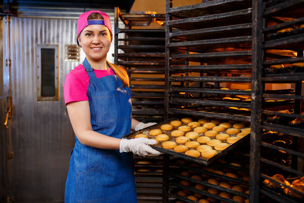 Una panadera profesional sostiene una bandeja con galletas frescas en sus manos. Pasteles dulces en una panadería - Foto, Imagen