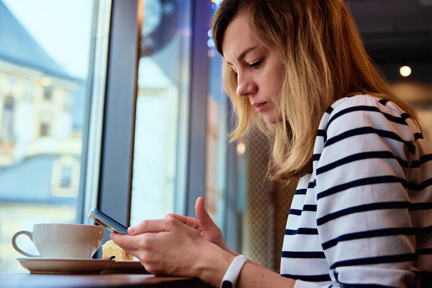 女性の手はカフェでスマートフォンを使用し、インターネットブラウザをサーフィンし、コーヒーブレイクを持っています。オンラインコミュニケーションとソーシャルメディア - 写真・画像